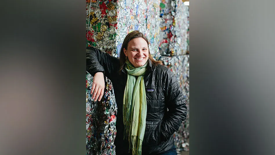 A smiling woman in a puffer coat and scarf leans against bales of aluminum cans
