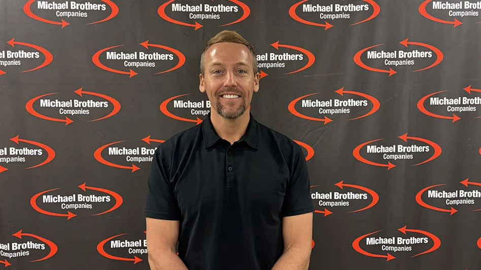 a smiling man with a beard before a micheal bros. step and repeat