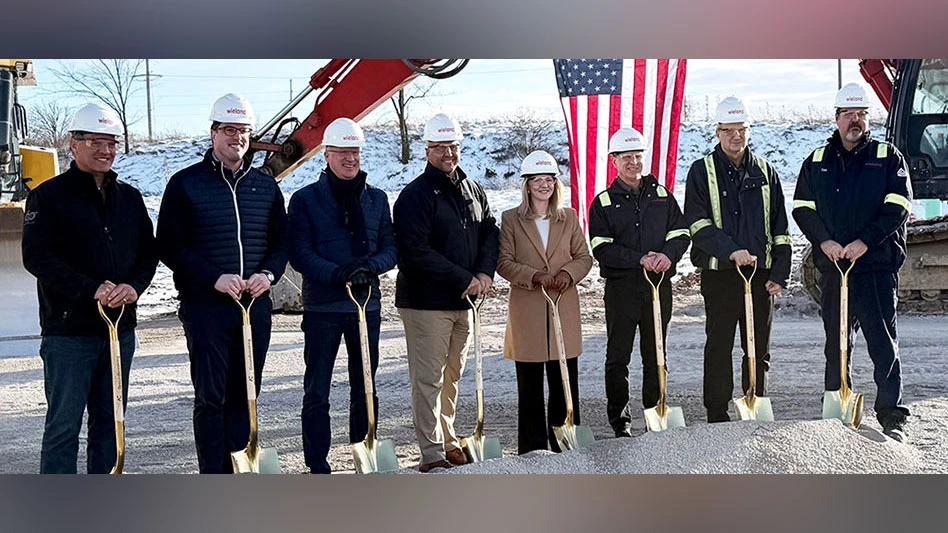 people wear hard hats and stand with shovels with heavy equipment and a u.s. flag behind them.